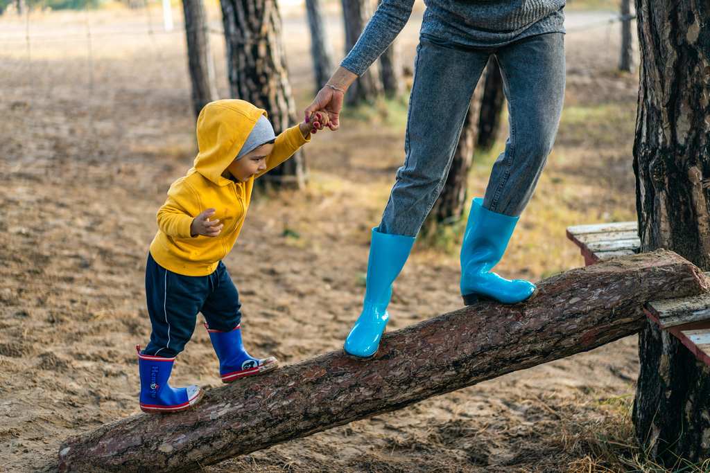 Bebê dando a mãozinha para um adulto e caminhando com ele sobre um tronco, ilustrando como incentivas o bebê a andar