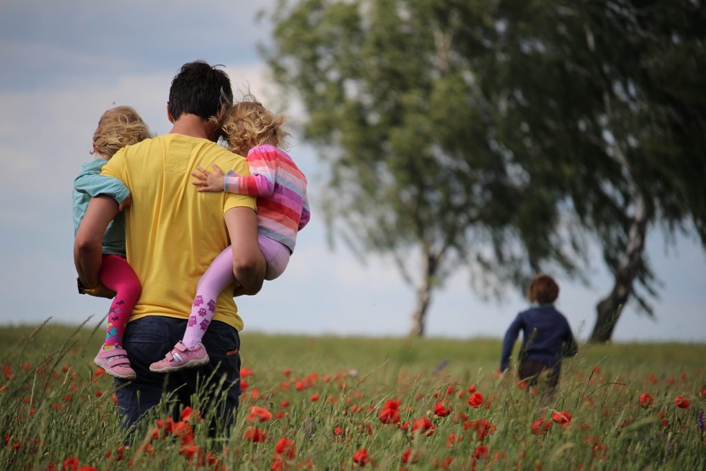 Em um ambiente externo,com grama, flores e duas árvores, um pai segurando duas crianças no colo, enquanto outra corre na frente. Simbolizando a importância das atividades psicomotoras e a família