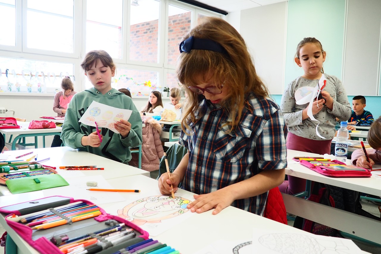 Crianças em sala de aula, uma menina está colorindo um desenho e outras duas estão recortando papéis com desenhos coloridos, simbolizando desenvolvimento neuropsicomotor e acompanhamento escolar