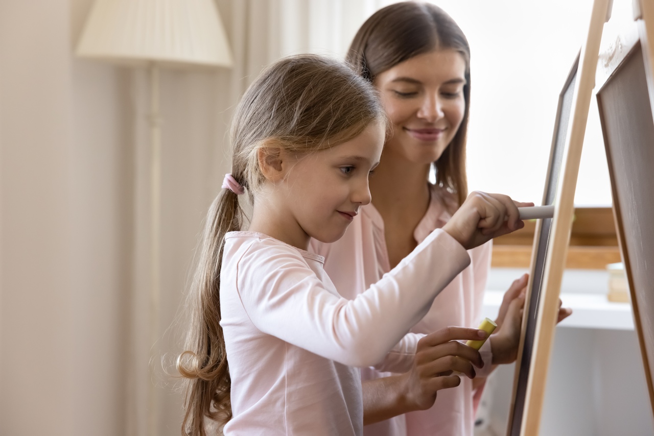 Simbolizando desenvolvimento neuropsicomotor: Menina criança sorrindo desenhando com giz em lousa, acompanhada da mãe sorrindo que também segura um giz; estão num ambiente com luz natural vinda da janela e uma luminária de chão apagada ao fundo